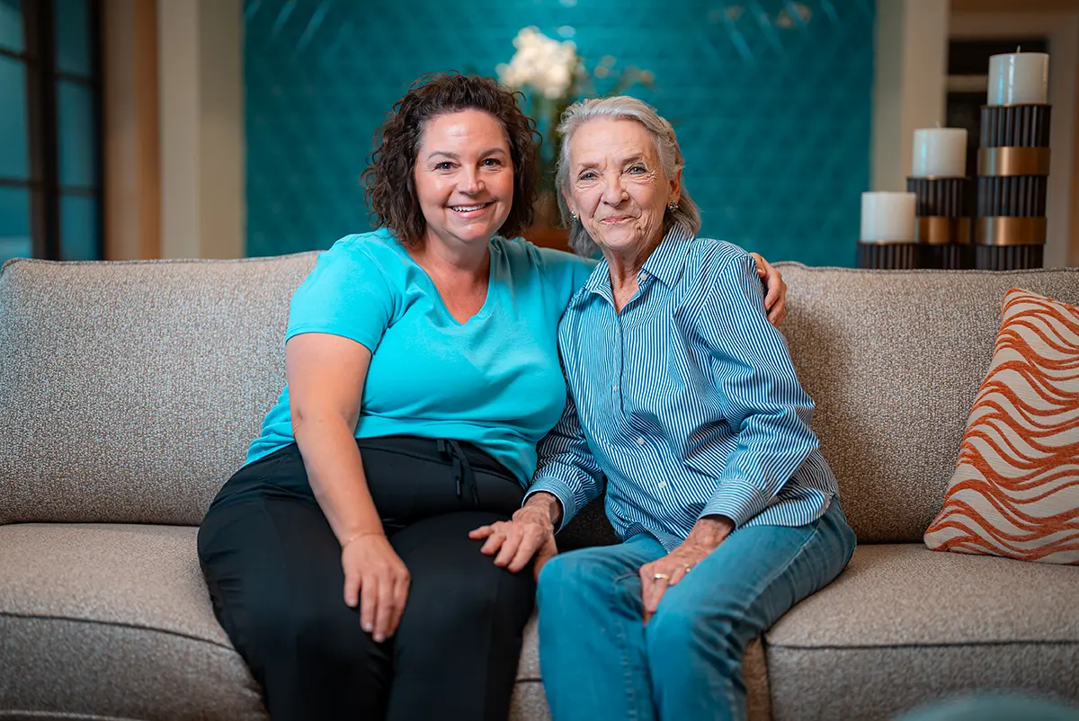 A senior adult and her adult daughter sit on a couch